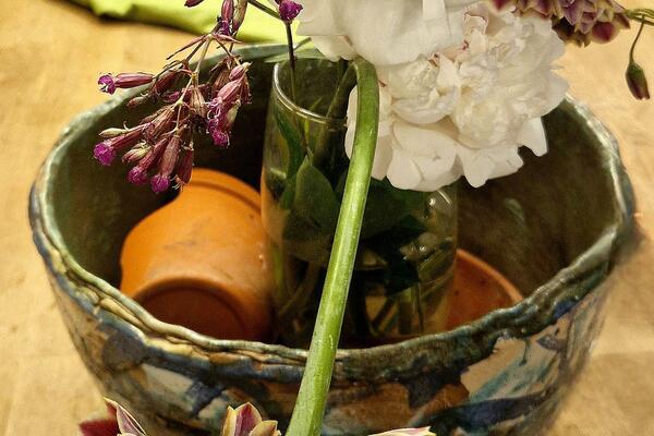 Blumenarrangement mit weißen Blüten in einer Glasvase innerhalb einer keramischen Schale auf einem Holztisch. Im Vordergrund liegen rosafarbene Blütenstiele, im Hintergrund sind Kleidungsstücke unscharf zu sehen.